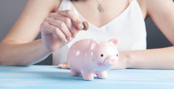 Pink piggy bank sat on a blue table with a woman holding a coin above to put in. Side income ideas for extra income - The Tonic