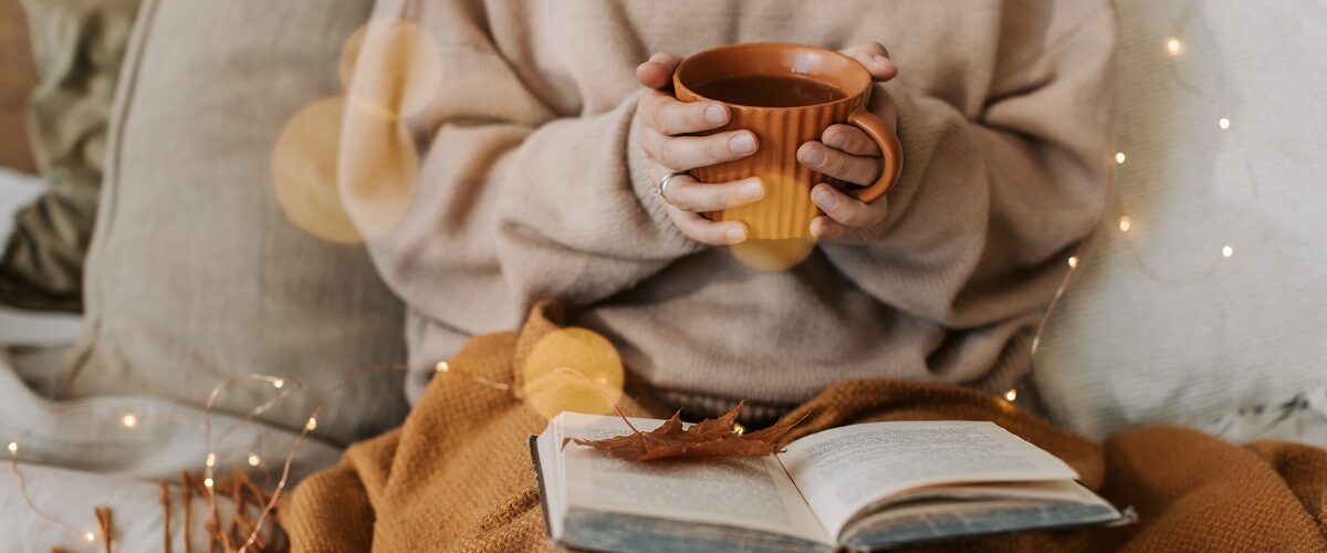 Image shows someone cosied up in blankets holding a cup for something warm to drink