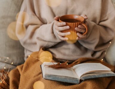Image shows someone cosied up in blankets holding a cup for something warm to drink