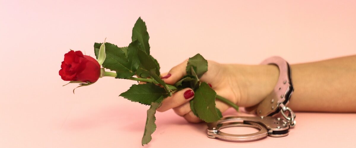 Hand reaching whilst holding a rose, with handcuff around wrist. Dating in cuffing season
