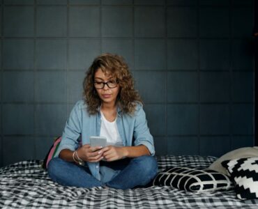 Image of a woman sat on a bed, looking down at her phone. She does not appear to be happy.