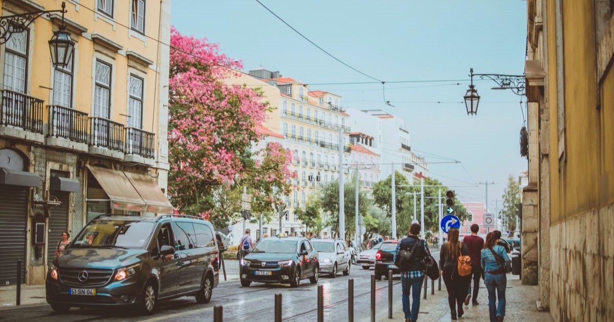 Image of the streets of Lisbon, with beautiful pale buildings, a pink tree, and a few people wandering down the street.